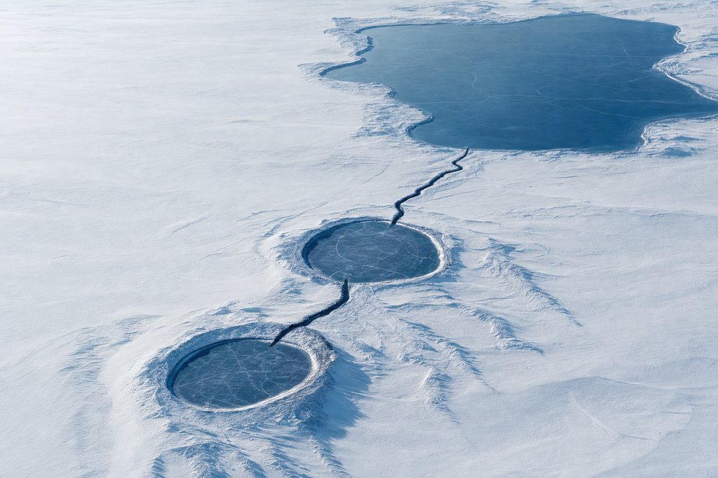 Aerial view of two round frozen lakes on a wide snow plain joined by one straight dark channel, with a third much larger frozen lake beyond them reached by a second channel — the plate for the Alberta escalation chain, which reaches a regulator
