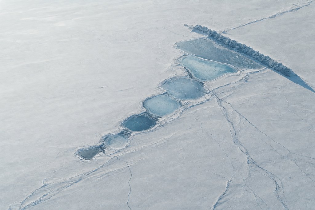 Aerial view of a chain of small shallow frozen ponds across a flat snow plain, the last one sealed off by a long raised pressure ridge — the plate for where the complaint chain stops in this province