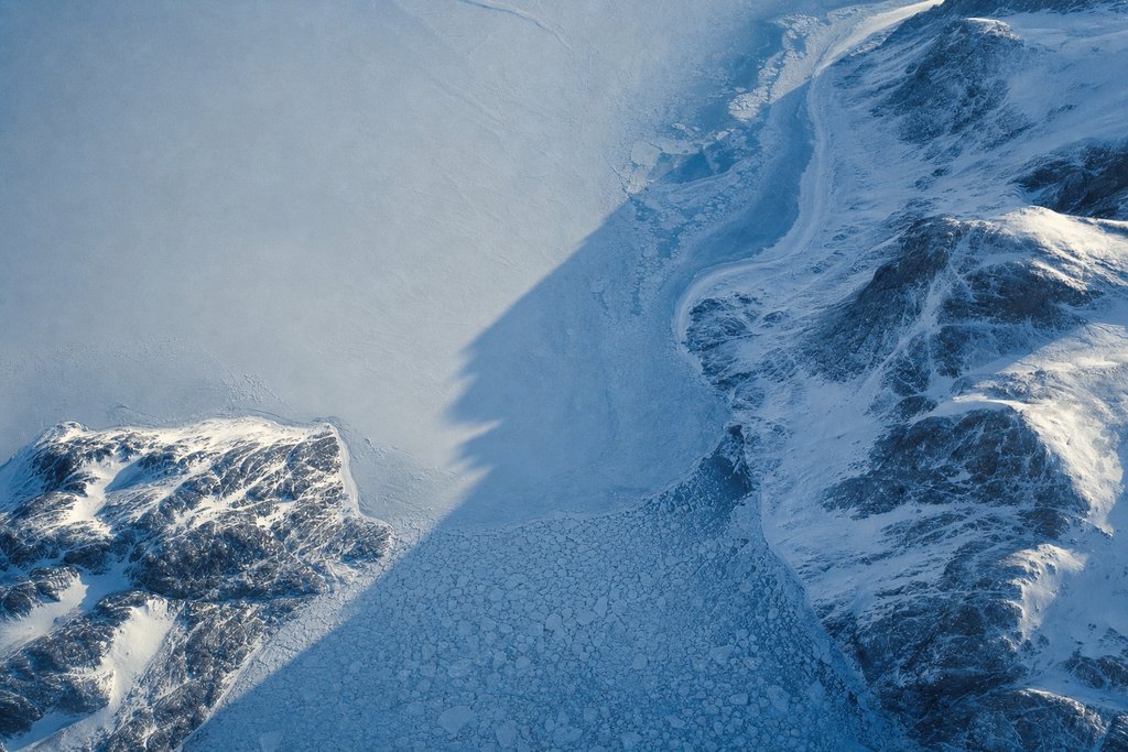 Aerial view of a tidal channel between two snow-covered rocky headlands, closed across its mouth by a solid bar of packed sea ice — the plate for where the complaint chain stops in this province