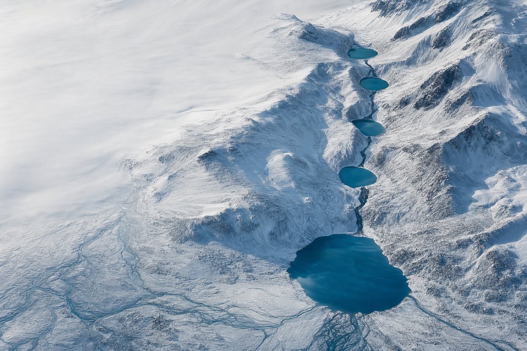 Aerial view of four frozen tarns descending a long snow-covered slope in a stepped chain, each linked to the next by a clear dark channel, the lowest and largest opening onto a broad plain — the plate for the Ontario escalation chain, which reaches a regulator