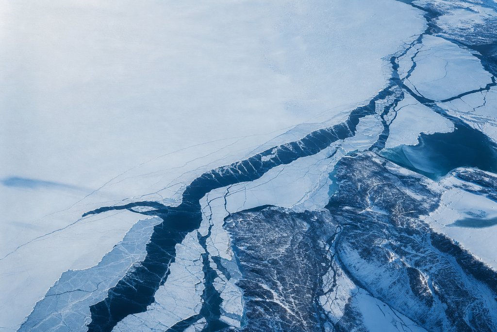 Close aerial view of lake ice breaking open, dark leads and fractures spreading across pale ice in branching channels — the plate for the payouts section