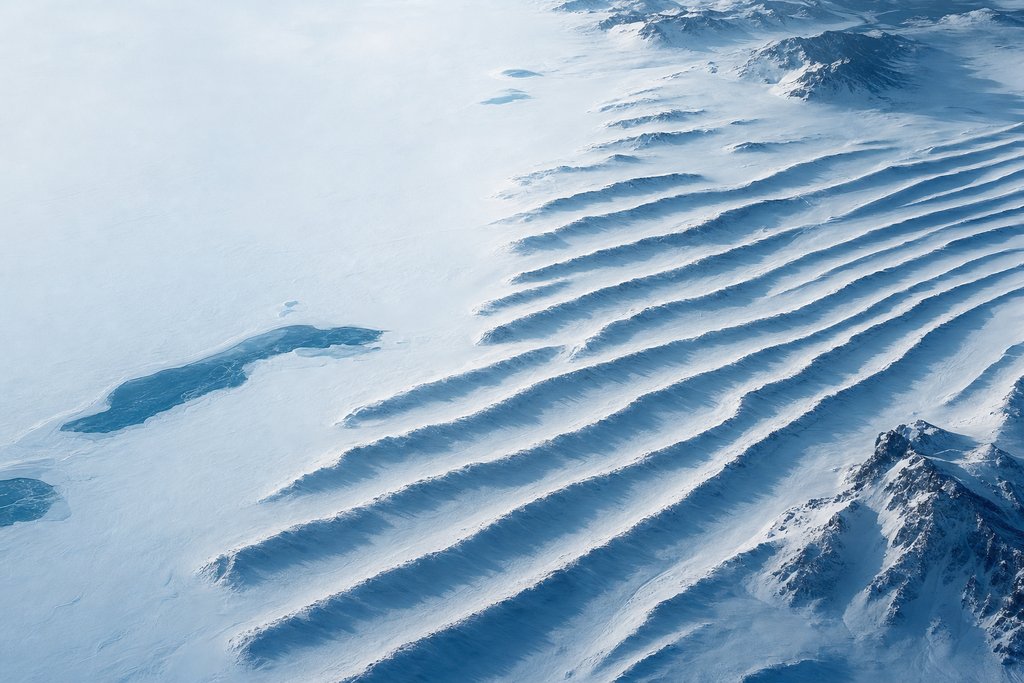 Aerial view of a wide snowfield crossed by a series of evenly spaced parallel natural drift ridges, each casting a long low shadow — the plate for about page