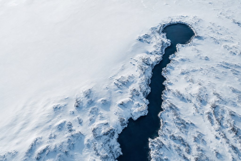 Aerial view of a narrow dark channel of open water leading inward from a snow-covered coast to one small sheltered circular basin — the plate for contact page