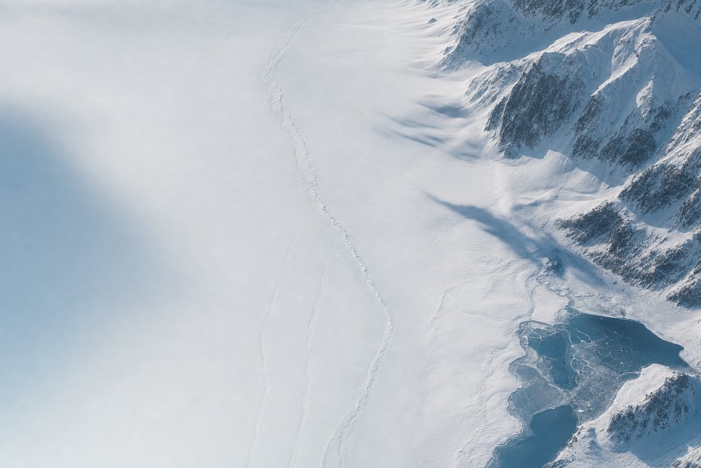 Aerial view of an old snow surface crossed by a long healed crack, its earlier line still faintly visible as a pale ridge beneath newer snow — the plate for corrections page