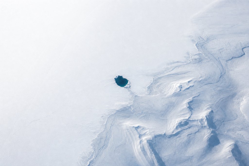 Aerial view of a single small dark opening in an otherwise unbroken white ice sheet, clear deep water visible through it — the plate for verify a certificate in the game fairness section