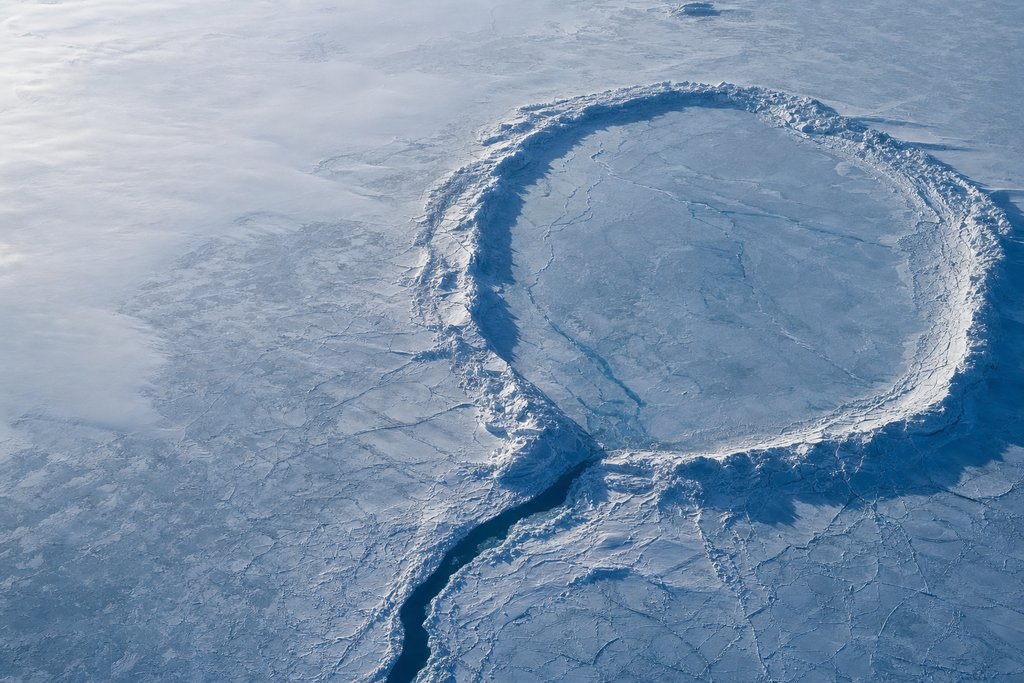 Aerial view of a large frozen lake enclosed by an unbroken raised ridge of pressure ice, with one clear dark channel leading out of it — the plate for the iGaming Ontario page