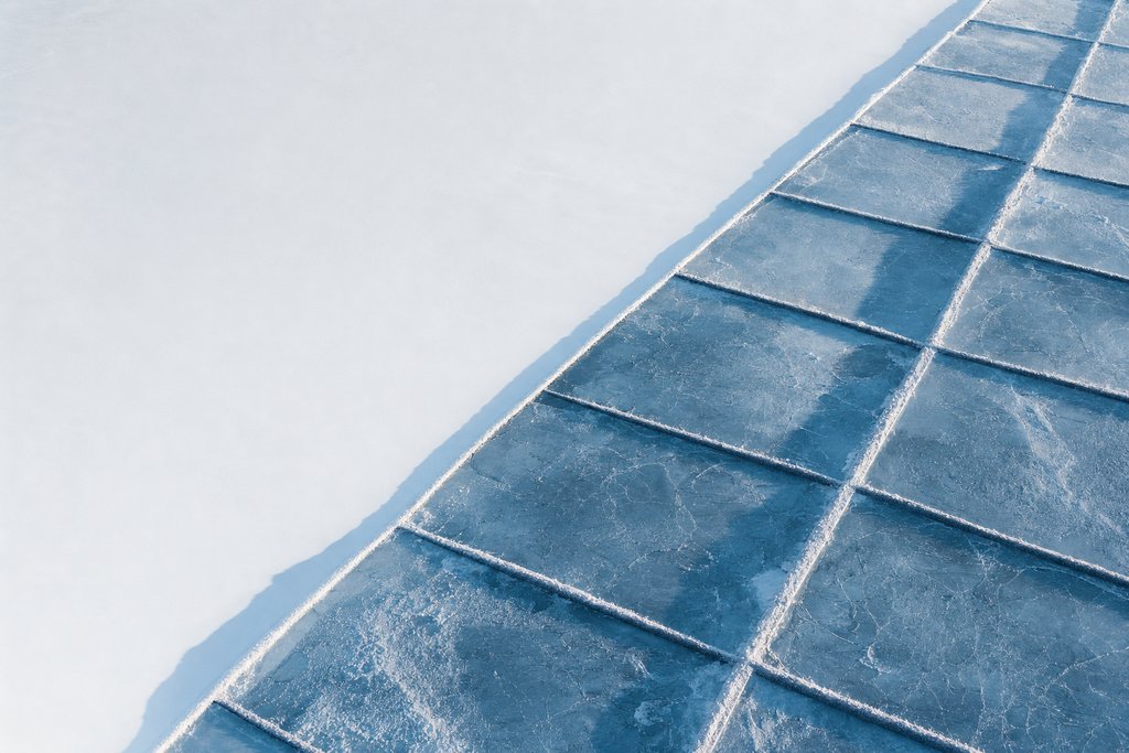 Aerial view of a precisely geometric field of rectangular ice panels filling one half of the frame and ending at a hard straight edge, beyond which the snow is entirely unmarked — the plate for malta mga in the licences section