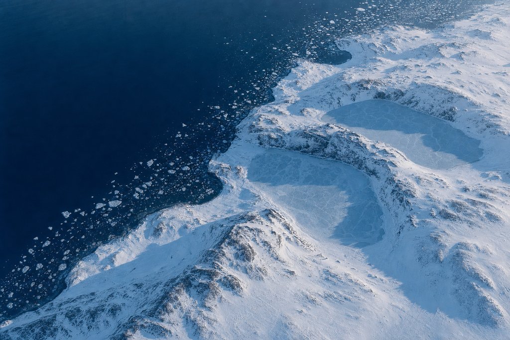Aerial view of a snow-covered Atlantic coastline with three separate frozen inlets and drifting pack ice offshore — the plate for Atlantic Lottery, the provincial site scored on the same rubric