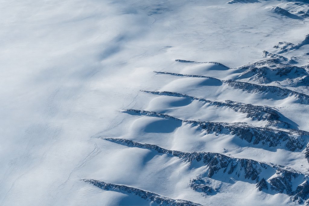Aerial view of a snowfield crossed by a series of low parallel drift barriers, each holding a bank of drifted snow behind it — the plate for deposit and loss limits in the safer gambling section
