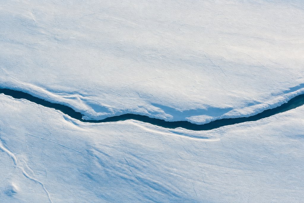 Aerial view of a vast featureless snow plain crossed by a single dark meltwater channel, the only feature in the frame — the plate for how every figure here is verified