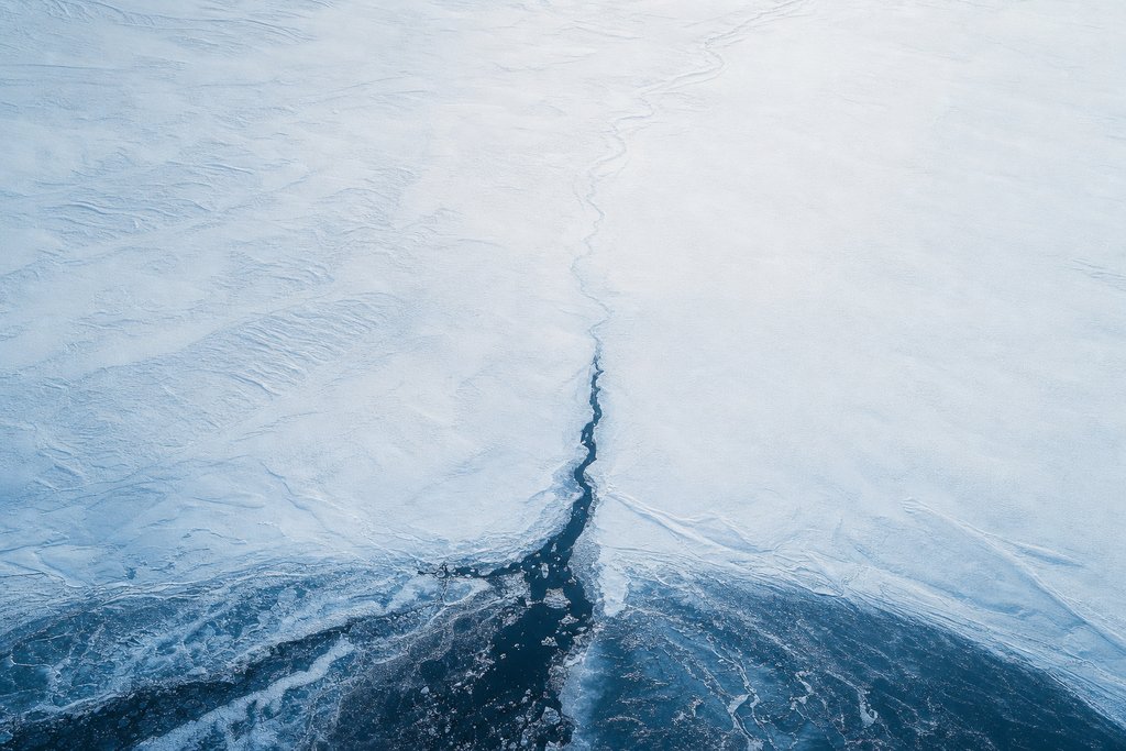 Aerial view of a dark water channel narrowing and disappearing beneath an unbroken sheet of snow, its continuing course visible only as a faint trace — the plate for what happens after you click withdraw