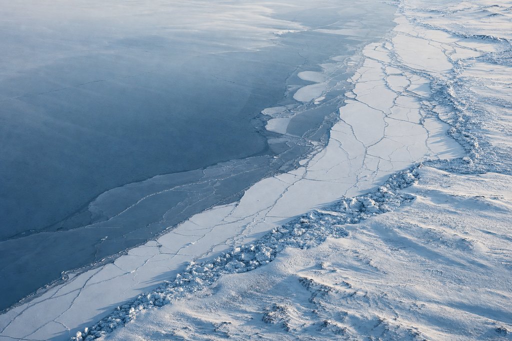 Aerial view of the frozen edge of a vast shallow lake, the ice cracked into long pale plates and pressure ridges — the plate for the Manitoba safety index