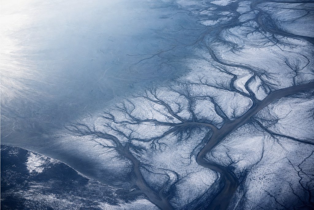 Aerial view of a wide tidal estuary at low tide in winter, sinuous drainage channels carved across pale frozen mudflats — the plate for the New Brunswick safety index