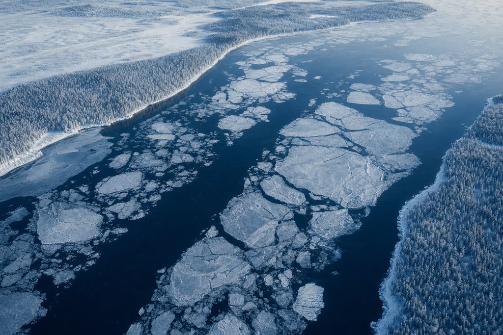 Aerial view of a wide frozen river channel with pale drifting ice pans on dark water, snow-covered boreal forest along both banks — the plate for the Quebec safety index