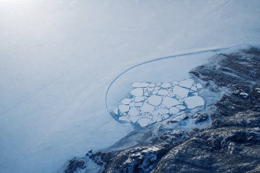 Aerial view of a large frozen lake with a small tight cluster of distinct pale ice plates near one shore enclosed by a single curved pressure ridge, and a vast empty expanse of unbroken ice beyond it — the plate for the Alberta register reading